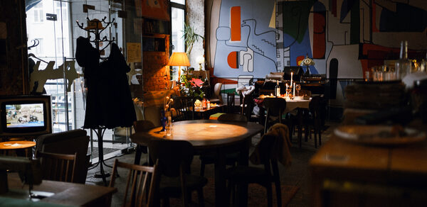 Гастробар Дневник. Зал с деревянными столами и стульями, пол из темного дерева. На стене абстрактная роспись. Слева стеклянная дверь с вешалкой для одежды. Освещение теплое, настольная лампа и свечи. В углу растения и книги на полках.