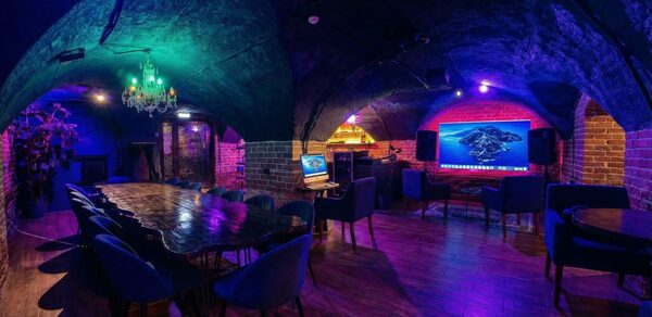 GB Rooms(ГБ Румс). Лофт №1. Подвальное помещение с арочными кирпичными стенами и потолками. Длинный деревянный стол с синими стульями. На стене большой экран, по бокам колонки. Декоративная люстра. Пол деревянный, освещение разноцветное. Компьютерный монитор на столе.