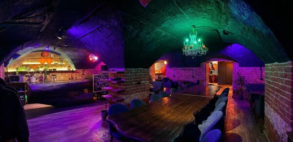 GB Rooms(ГБ Румс). Лофт №1. Кирпичное подвальное помещение с арочным потолком. Длинный деревянный стол, окружённый стульями. Барная стойка на заднем плане. Зелёная и фиолетовая подсветка. Классическая люстра. Пол деревянный. Мягкая мебель вдоль стен.