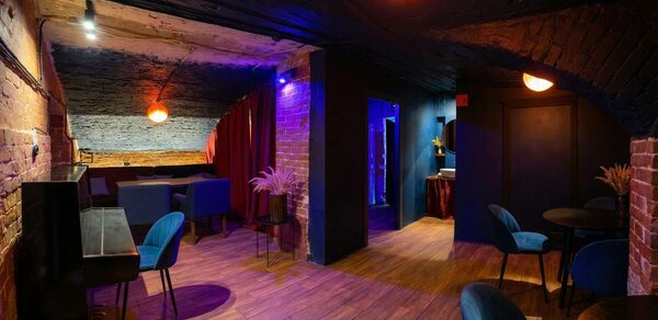 GB Rooms(ГБ Румс). Лофт №1. Подвальное помещение с кирпичными стенами и деревянным полом. Зоны с мягкими синими креслами и диванами, столики. Яркое цветное освещение, красные шторы. Потолок темного цвета. Круглые лампы с теплым светом.