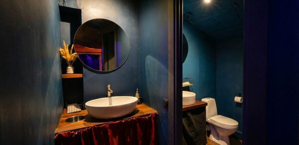 GB Rooms(ГБ Румс). Лофт №1. Ванная комната с синими стенами, круглым зеркалом над раковиной, круглой раковиной на деревянной столешнице, унитазом. Металлический дозатор мыла, красная занавеска под столешницей. Полки с декором, верхнее точечное освещение.