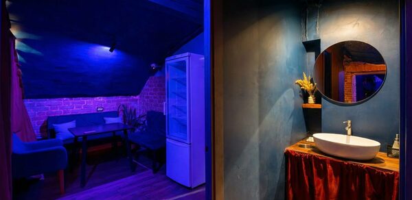 GB Rooms(ГБ Румс). Лофт №1. Комната с синим и красным освещением, кирпичные стены, черная мебель, обеденный стол, диваны, холодильник, круглая раковина с зеркалом, деревянный и керамический декор, занавеска красного цвета, темный деревянный пол.