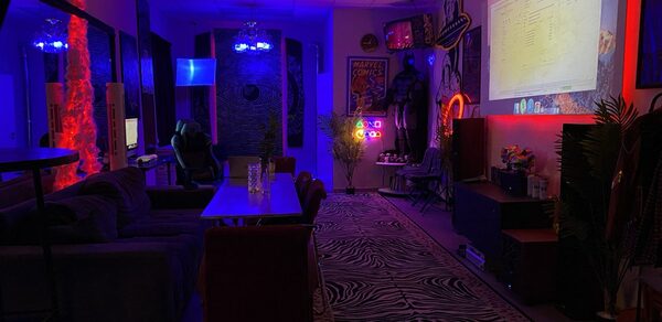 GB Rooms(ГБ Румс). Лофт №2. Комната с диванами и креслами, прямоугольный стол, проектор с экраном. Яркое разноцветное освещение, неоновые вывески, настенные постеры. Ковёр с зебровым узором, растения в горшках. Потолочные светильники, мебель из тёмных материалов.