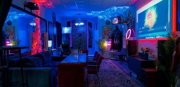 GB Rooms(ГБ Румс). Лофт №2. Квартира с затемнённым освещением, столом и креслами, диванами, неоновыми вывесками и декоративными элементами. На стенах постеры, есть крупный экран для проекций. Используются яркие синие и красные цветовые акценты. На полу ковер с узором.