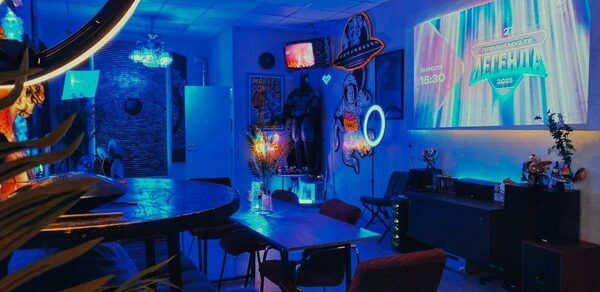 GB Rooms(ГБ Румс). Лофт №2. Комната с неоновым освещением, большой стол и стулья, диваны, проектор с изображением на стене, тематические постеры и декоративные элементы. Стены светлые, пол паркетный, на некоторых поверхностях светящиеся неоновые элементы.
