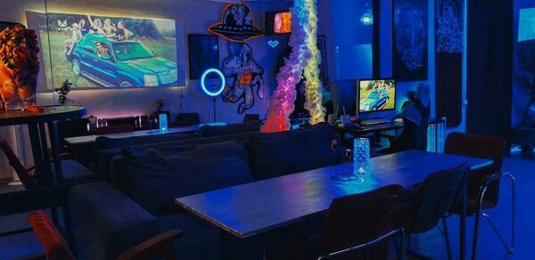 GB Rooms(ГБ Румс). Лофт №2. Помещение для отдыха с неоновым освещением в синих тонах. Длинный стол с несколькими стульями, мягкое сиденье, телевизор, настенные картины. Пламя в центре стола. Просторная обстановка с тёмной мебелью.