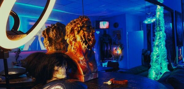 GB Rooms(ГБ Румс). Лофт №2. Комната с ярко-синим и фиолетовым освещением, декоративные бюсты, круглый стол с металлическими предметами, кольцевая лампа, диван, экран, декорированные стены, светящийся элемент в углу, модерн-стиль, открыт веер.