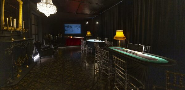 Лофт Космос. Зал Президент. Тёмная комната с двумя покерными столами и прозрачными стульями. На одной стене висят чёрные шторы, на другой — камин с зеркалом и подсвечниками. На потолке хрустальная люстра. В углу красный стол с проектором и экраном. Пол узорный, темный.