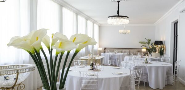 Лофт Космос. Зал Президент. Светлое банкетное пространство с белыми стенами и большими окнами, украшенными шторами. Круглые столы с белыми скатертями и прозрачными стульями. Потолочные люстры и настольные лампы создают освещение. В центре композиции белые каллы в вазе.