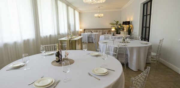 Лофт Космос. Зал Президент. Просторная комната с круглыми столами, накрытыми белыми скатертями; прозрачные стулья; столовые приборы и бокалы на столах; большая люстра на потолке; светлые шторы на окнах; растения в углу; нейтральные тона.