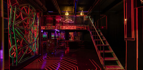 Лофт Космос. Зал Ночной Клуб. Темное помещение с черным потолком и стенами, разноуровневая планировка с лестницей. Неоновая подсветка красного и синего цветов на полу и стенах создает геометрические узоры. Металлические конструкции, подвесные лампы.
