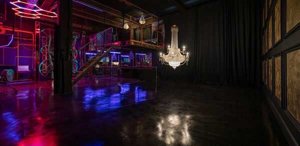 Лофт Космос. Зал Ночной Клуб. Просторное помещение с бетонным полом и черными стенами. Лестница с прозрачными перилами ведет на второй уровень. Неоновая подсветка, люстра и несколько висящих ламп освещают пространство. Часть стены закрыта плотной черной занавеской.