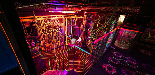Лофт Космос. Зал Ночной Клуб. Креативное пространство с ярким неоновым освещением и геометрическими узорами. Металлические конструкции и лестница вниз, разноцветные световые линии на стенах и потолке, стеклянные панели. Тёмный фон, акцент на фиолетовый и оранжевый цвета.