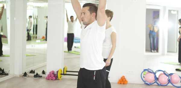 Face & Body(Лицо и Тело). Фитнес-зал с зеркалами на стенах, деревянным полом. В зале присутствуют коврики для йоги, гантели, гимнастические шары. Освещение яркое, потолочные светильники. Люди выполняют упражнения.