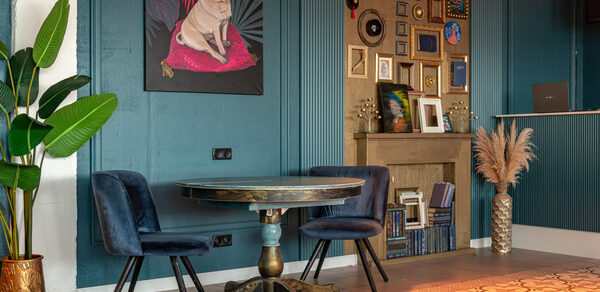 Лофт Май. Стильное пространство с верандой. Угловое пространство с деревянным круглым столом и двумя синими стульями. На стене синие панели, висят картины и фотографии. Полка с декором и несколькими книжными полками. На полу плитка с узором, рядом растение в горшке.
