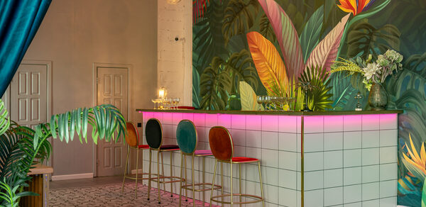 Лофт Май. Стильное пространство с верандой. Барная стойка с белыми квадратными плитками, подсветка розового цвета. Три барных стула с разноцветными спинками. Стена с ярким тропическим рисунком. Зона декорирована зелеными растениями, висят круглые подвесные светильники.