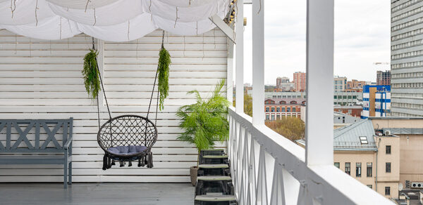 Лофт Май. Стильное пространство с верандой. Терраса с белыми деревянными стенами и крышей, пол серый. Плетеное подвесное кресло, рядом скамейка и табуреты. Растения в горшках. Перила с крестовинами. На заднем плане вид на городскую архитектуру.
