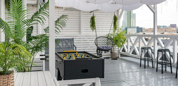 Лофт Май. Стильное пространство с верандой. Открытая терраса с серым деревянным полом, белыми стенами и тканевым потолком. Мебель: диваны, стулья, столики, футбольный стол. Растения в горшках. Вид на город.