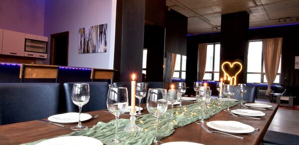 Элегантный лофт с хорошим звуком и балконом. Столовая зона с длинным деревянным столом, сервированным стеклянными бокалами и тарелками. В центре стола зеленая ткань и свечи. Мягкие стулья вдоль стола. На стене картина, сбоку кухня. Светодиодное освещение фиолетового цвета.