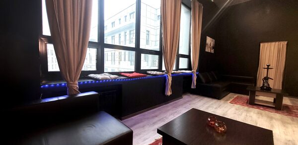 Элегантный лофт с хорошим звуком и балконом. Лофт с чёрными стенами и большими окнами, с бежевыми шторами. Низкие чёрные кожаные диваны и деревянные столы. Красные ковры на светлом полу. На одном из столов расположен кальян. Искусственное освещение, вид на город.