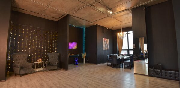 Элегантный лофт с хорошим звуком и балконом. Просторное помещение с высокими потолками и деревянным полом. Черные стены, на одной из которых гирлянда. Зона с креслами и столиком, рядом телевизор. Обеденная зона с большим столом и стульями. Потолочные светильники и люстра. Большие окна с занавесками.