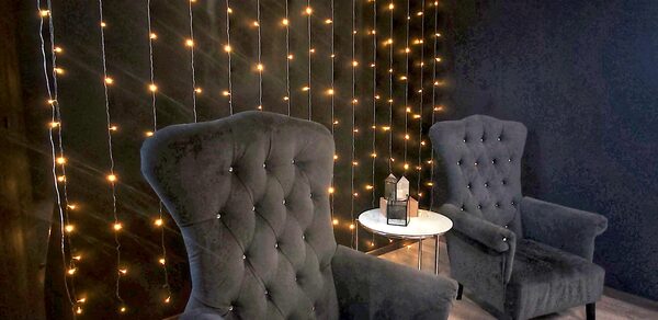 Элегантный лофт с хорошим звуком и балконом. Тёмно-серая комната с двумя мягкими креслами, круглым белым столиком, на котором стоит декоративный светильник. На стене гирлянда из маленьких жёлтых огней. Пол из светлого дерева.