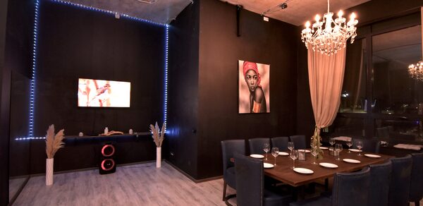 Элегантный лофт с хорошим звуком и балконом. Пространство для мероприятий с деревянным столом и синими стульями, сервированное столовыми приборами и бокалами. Черные стены, подсветка по периметру одной стены, телевизор, картина. Люстра с лампами, светлая штора, декоративные элементы.