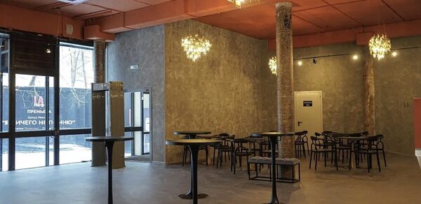 Театр в Хамовниках. Просторное помещение с высокими потолками, бетонные стены и пол, оранжевый потолок. Столики с высокими черными стойками и черными стульями. Большие окна, несколько круглых люстр. Прозрачные стеклянные двери, виден уличный баннер.