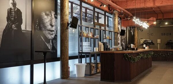 Театр в Хамовниках. Барная зона в просторном помещении с высокими потолками. Стойка из темного дерева с растительной декорацией. Стены украшены крупными черно-белыми портретами. Освещение подвесное, имеется множество бутылок. Потолок красный.