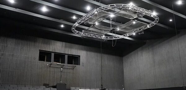 Театр в Хамовниках. Театральное или конференц-пространство с амфитеатром, оборудовано рядами серых кресел. Потолок черный с вмонтированными светильниками и подвесной конструкцией. Стены облицованы темным древесным материалом.