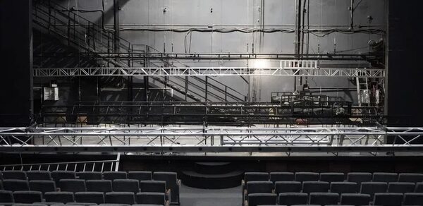 Театр в Хамовниках. Театральный зал с рядами серых кресел и сценой. На сцене установлены металлические конструкции и оборудование. Задняя стена сцены с проводами и аппаратурой. Освещение верхнее, стены серые.