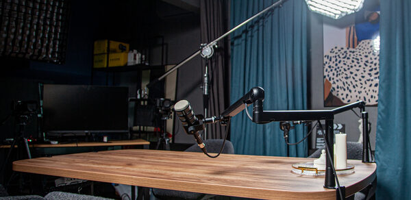 Студия для записи подкастов "Искусство Подкаста". Студия с деревянным столом, оборудованная микрофонами на стойках. Освещение достигается за счет большого софтбокса. На заднем плане шторы и декоративное панно. В помещении также есть стеллажи с техникой и экран.