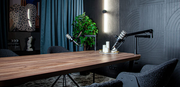 Студия для записи подкастов "Искусство Подкаста". Современное помещение с деревянным столом, вокруг которого расположены мягкие стулья. На столе микрофоны на стойках, декор в виде свечей и зелёного растения. Стена с абстрактными панелями и вертикальным светильником, тяжелые синие шторы.