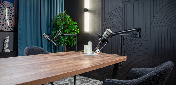 Студия для записи подкастов "Искусство Подкаста". Комната со столом из дерева, оборудованным микрофонами. Светильники на черно-текстурной стене. Серые стулья, синий занавес и зеленое растение. На полу ковёр светлого цвета.