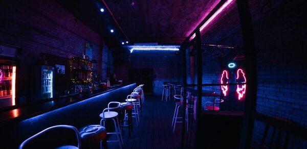PVZ CLUB(ПВЗ Клуб). Бар с длинной стойкой, за которой стоят высокие стулья. Освещение неоновое, синие и розовые оттенки. На стене рисунок неонового ангельского крыла. Обстановка из тёмного кирпича, есть холодильник и полки с бутылками.