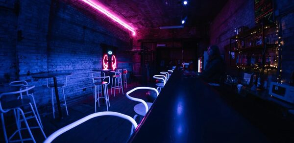 PVZ CLUB(ПВЗ Клуб). Барное пространство с высоким потолком и кирпичными стенами, обставлено высокими столами и стульями. Неоновая подсветка фиолетового цвета, барная стойка справа. На стенах полки с напитками, справа заметны гирлянды.