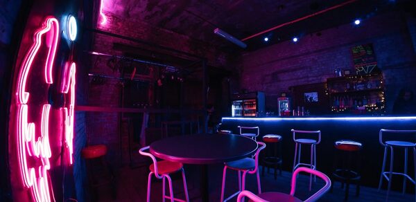 PVZ CLUB(ПВЗ Клуб). Тёмное помещение с барной стойкой, окруженной высокими стульями с красными сиденьями. Справа неоновые огни и вывеска на стене. Кирпичные стены, деревянный пол. Освещение синих и фиолетовых оттенков. На барной стойке видны напитки и декорации.