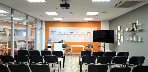 Конференц-зал. Конференц-зал с креслами на несколько рядов, бело-оранжевая стена с надписями, большая плоская панель на подставке, стеклянная перегородка слева, светлые стены и пол, потолочные светильники, кондиционер, полки с наградами и сертификатами.