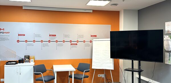 Конференц-зал. Конференц-зал с оранжевой акцентной стеной и инфографикой. Телевизор на стойке, флипчарт, два серых кресла и белый стол. Белый шкаф слева, напольное покрытие из светлого ламината. Яркое освещение.
