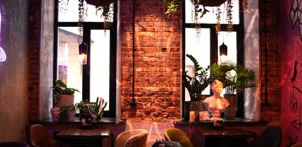 Винный бар “Дело не в вине”. Лофт-пространство с кирпичными стенами и высокими окнами. Разнообразные кресла и столы, декоративные растения и бюст. Подвесные светильники обеспечивают мягкое освещение. Яркий, уютный интерьер с граффити на стенах и неоновыми акцентами.