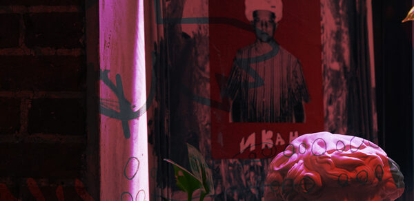 Винный бар “Дело не в вине”. Темное арт-пространство с кирпичной стеной, картина в рамке с изображением человека в шляпе на красном фоне, гипсовая скульптура и растение на переднем плане, цветное освещение.