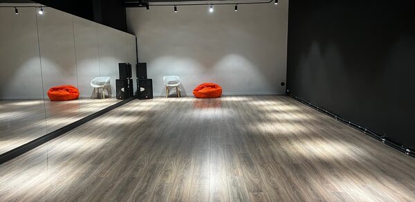 The Flow Space(Пространство Поток). Танцевальная студия с зеркальной стеной, деревянным полом, черно-белыми стенами. Две оранжевые мягкие подушки и два белых стула вдоль стены. Навесные черные светильники освещают пространство. Акустические колонны у стены.