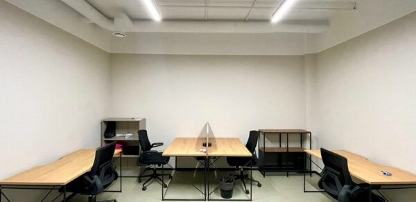 Smart Coworking(Умный Коворкинг). Офисное пространство с пятью рабочими местами, каждое с деревянным столом и черным офисным креслом. Белые стены, белый потолок с трубами и подвесным длинным освещением. Пол светлый, на столах и полу несколько предметов.