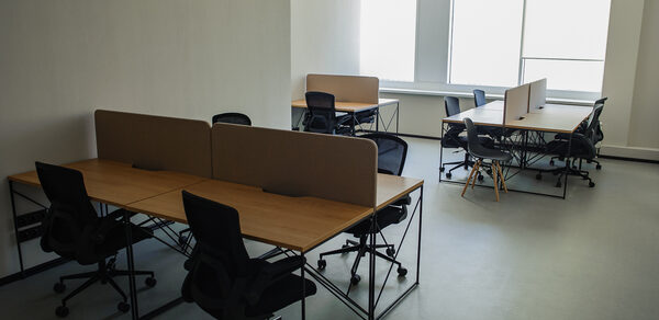 Smart Coworking(Умный Коворкинг). Офисное помещение с несколькими рабочими столами и офисными креслами. Столы разделены перегородками. Светлые стены и пол. Большие окна обеспечивают естественное освещение. Черная мебель с деревянными столешницами.