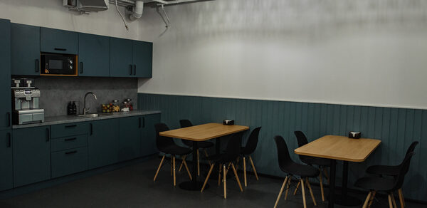 Smart Coworking(Умный Коворкинг). Кухонное помещение с темными шкафами, встроенной микроволновкой, кофемашиной и мойкой на кухонной стойке. Две деревянные столешницы с черными стульями. Светильники на потолке, серый пол и стены с панелями.
