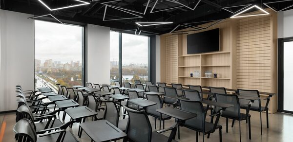 YES Technopark(ДА Технопарк). Конференц-зал. Конференц-зал с рядами черных стульев с пюпитрами, большие окна, современные геометрические светильники, полированные полы, деревянные панели на стене, экран с монитором, бутылки с водой на полке.