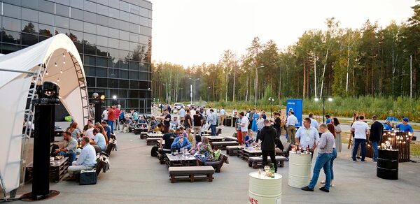 Технопарк «Университетский». Открытая площадка.. Открытое пространство с деревянными диванами и столами, белая тентовая конструкция, металлические бочки используются как высокие столы, панорамное стеклянное здание на заднем плане, вечернее освещение, зелёные деревья вокруг.