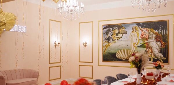 Барокко. Элегантный лофт. Праздничное пространство с длинным столом, покрытым белой скатертью и украшенным цветами. На стене объемная картина. Светильники на стенах и хрустальные люстры. Бежевые стены с золотыми элементами и неоновая вывеска. Красные и золотые шары на полу.