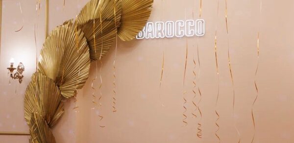 Барокко. Элегантный лофт. Бежевый интерьер с декоративной золотой аркой из листьев на стене, надписью из неоновых букв, светло-розовым диваном в углу. Потолочные золотые ленты спускаются вниз. Настенное бра с двумя лампами.