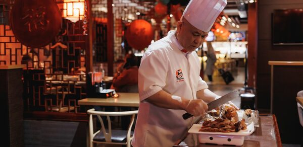 Ресторан J.Z.Peking Duck (J.Z. Утка по-пекински) . Ресторанное пространство, повар в форме режет мясо на передвижном столике. Слева решетчатая перегородка, декорированная красным кругом. Интерьер в тёплых тонах, видны красные и черные шары, приглушенное освещение, деревянная мебель.